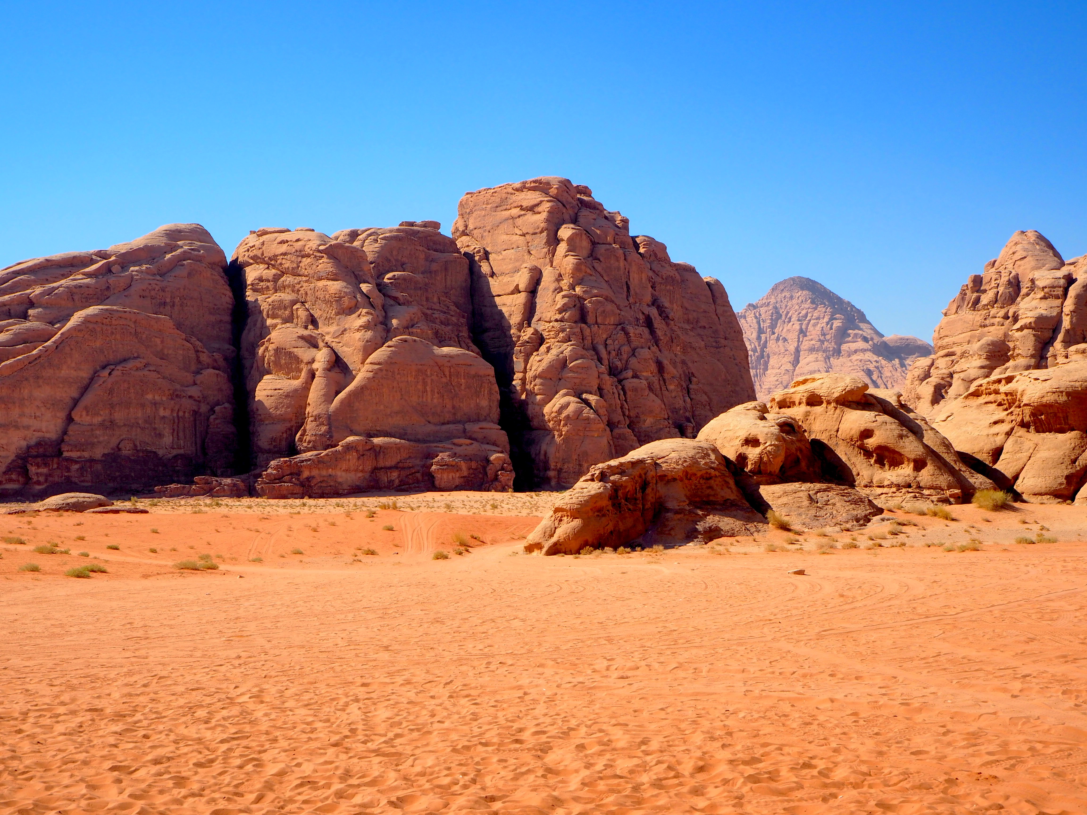 Towering sandstone rock formations rise above orange desert sand under a clear blue sky