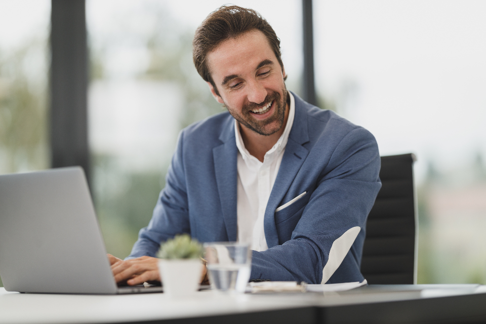 A smiling man in a blue blazer works on a laptop at an office desk